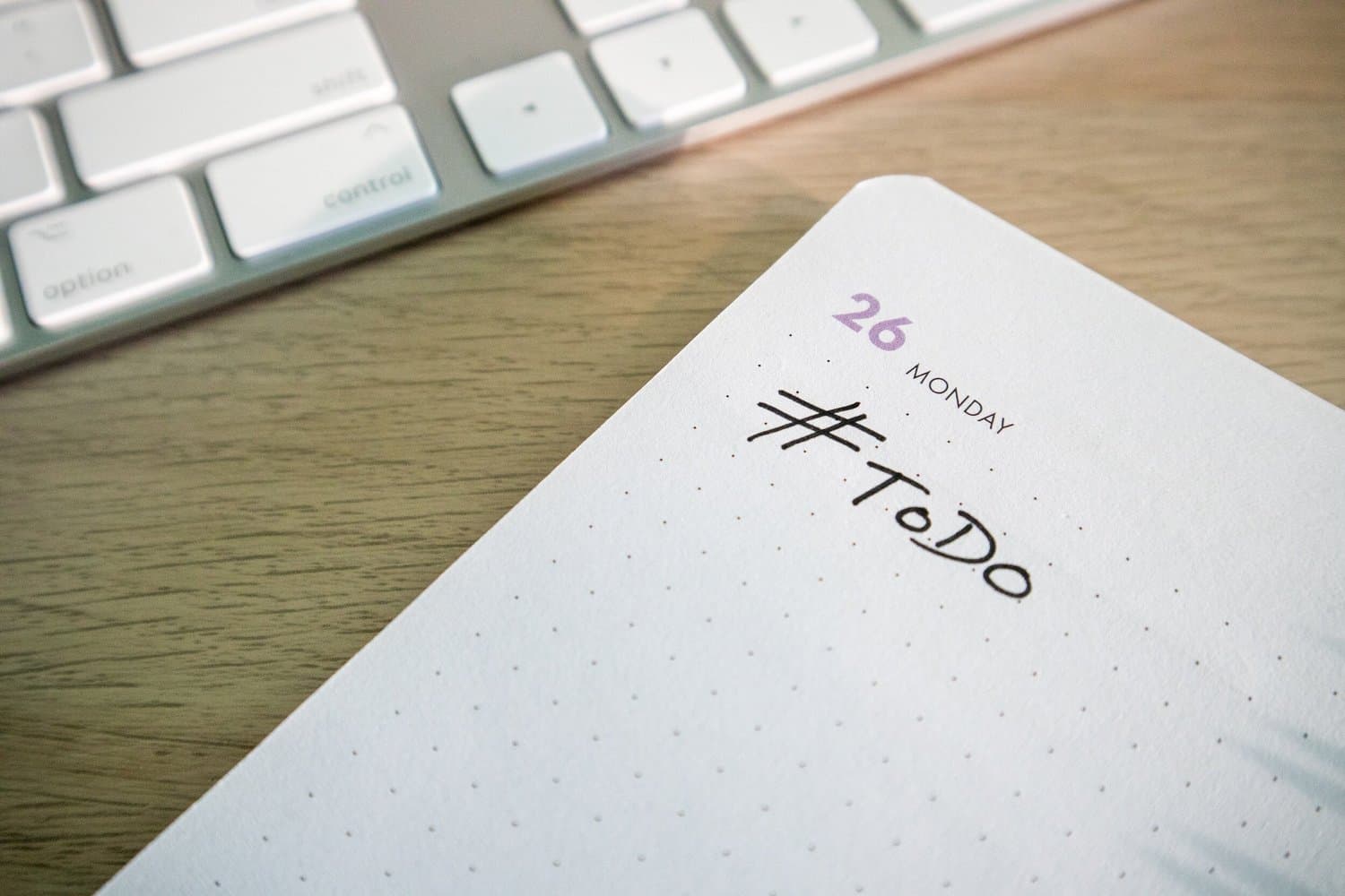 A close up photo of a white dot grid notebook on a wood desk beside a keyboard, open to a page headed 26 MONDAY with hashtag ToDo handwritten below.