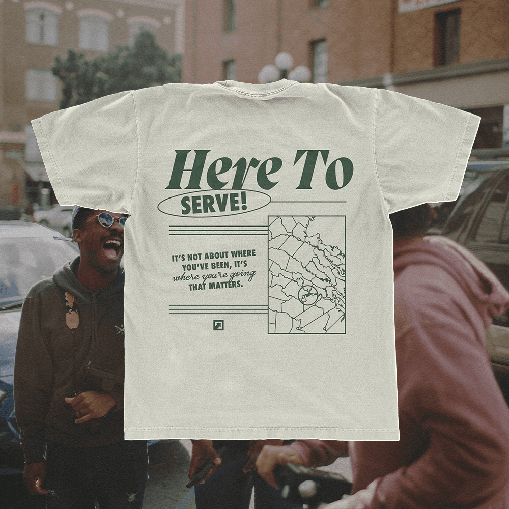 A merch mockup of a cream tee held up on a city street, printed Here To SERVE! in green serif beside a line drawn regional map and a short quote.