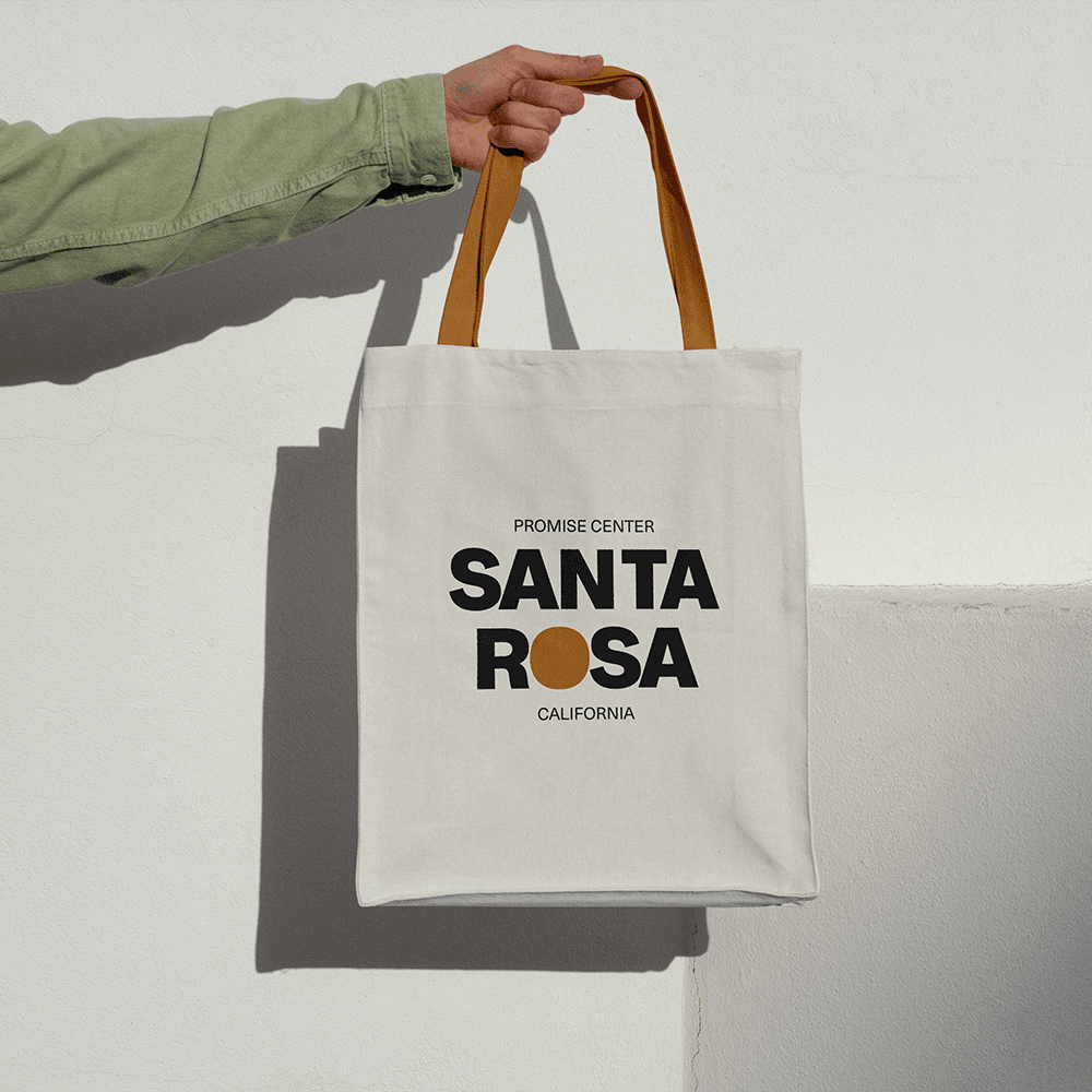 A natural canvas tote with tan handles held against a white wall, printed PROMISE CENTER SANTA ROSA CALIFORNIA in black capitals with an orange dot for the O.