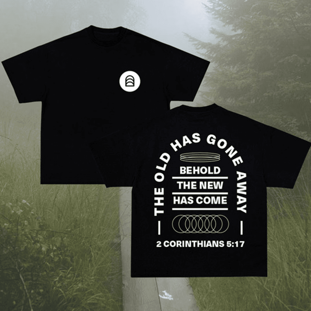 A merch mockup of two black tees in foggy woods, the back reading THE OLD HAS GONE AWAY BEHOLD THE NEW HAS COME in a white arch, 2 CORINTHIANS 5:17.
