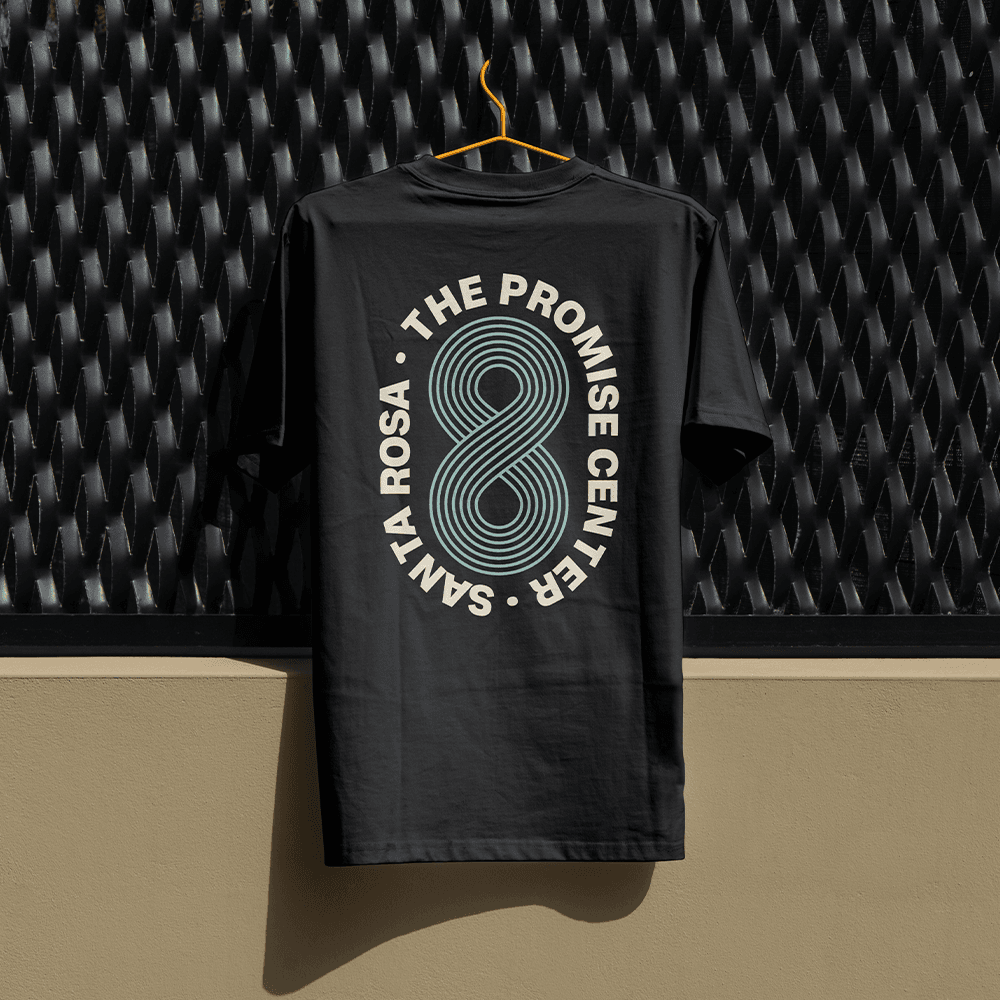 A black tee on an orange hanger against corrugated metal, printed THE PROMISE CENTER SANTA ROSA in a circle around a teal concentric figure eight.