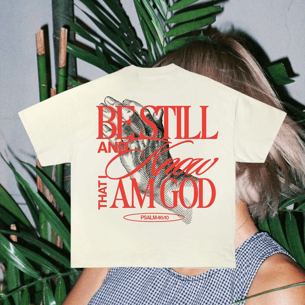 A merch mockup of a pale yellow tee worn among palms, printed BE STILL AND Know THAT I AM GOD in red over an engraved praying hands, marked PSALM 46:10.