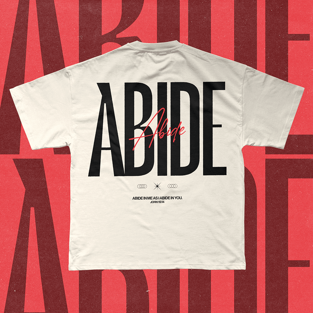 A merch mockup of a cream tee on a red backdrop, printed ABIDE in tall black capitals crossed by red script, with ABIDE IN ME AS I ABIDE IN YOU, JOHN 15:14.
