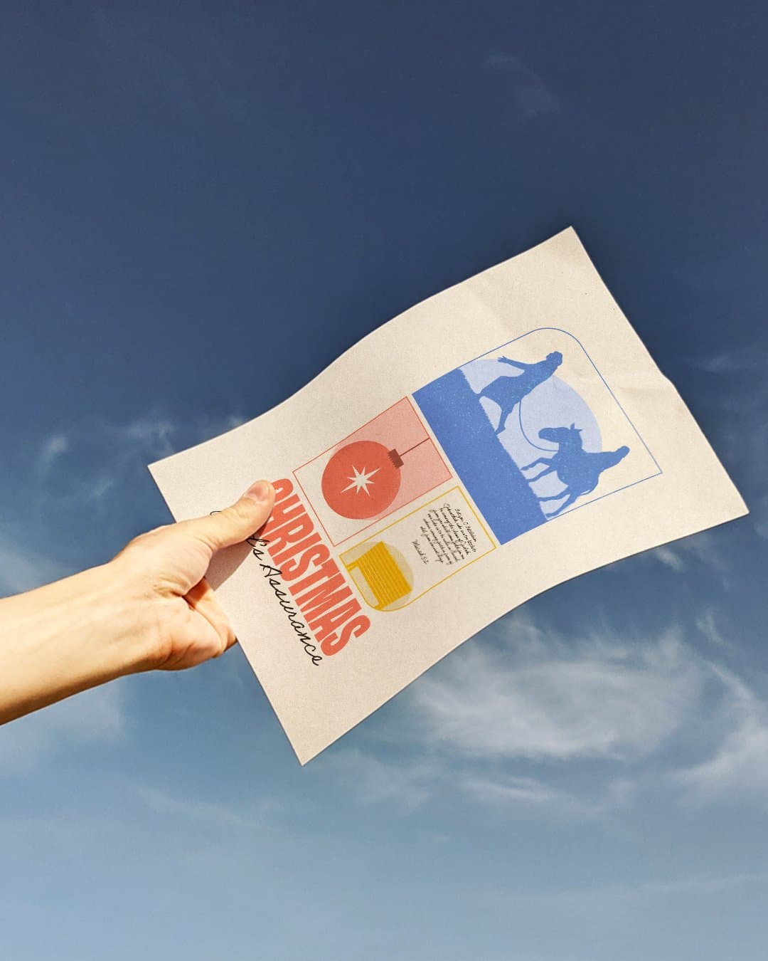 A hand holding the printed CHRISTMAS God's Assurance poster against a blue sky, showing the red ornament and blue donkey panels.