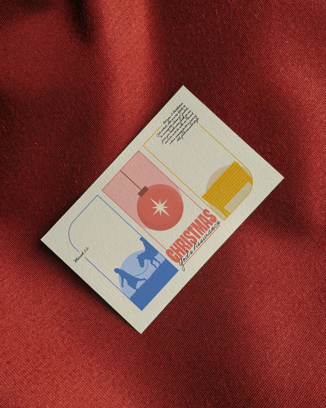 The CHRISTMAS God's Assurance card photographed at an angle on red fabric, showing the red ornament, yellow manger and blue nativity panels.