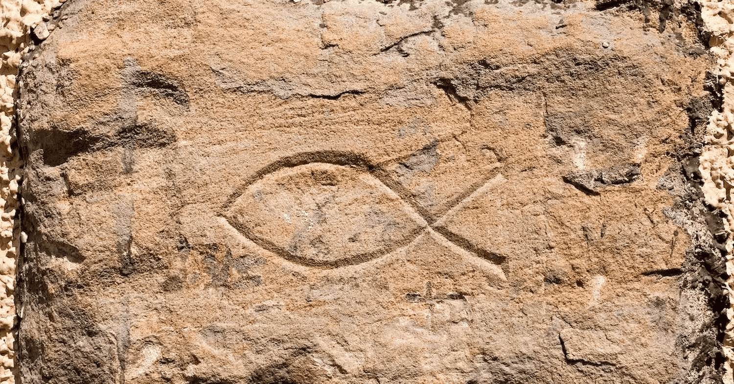 A close photograph of an ichthys fish symbol carved into a weathered sandstone block, its two shallow curved lines crossing at the tail.