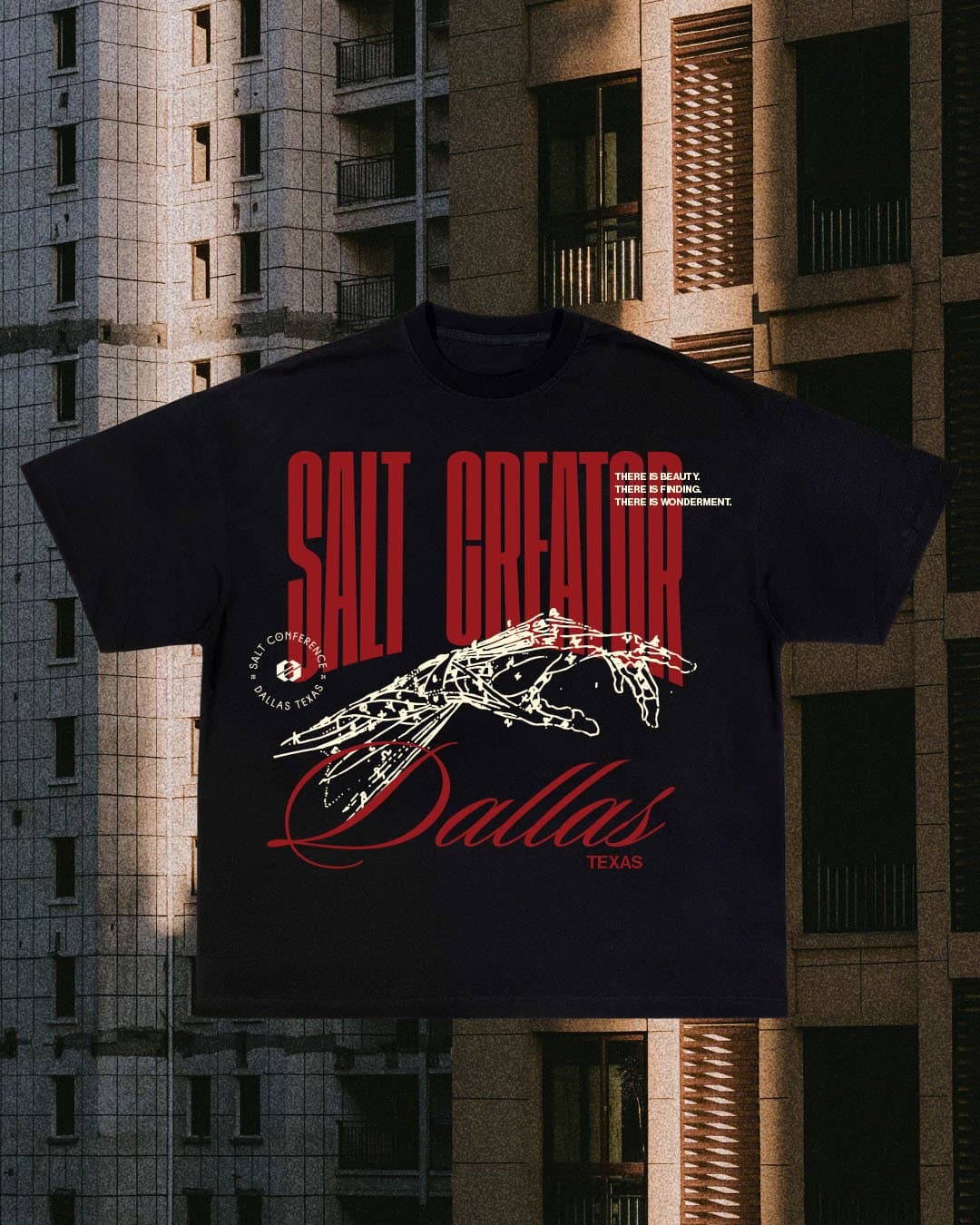 A merch mockup of a black tee against a concrete apartment block, printed SALT CREATOR in red capitals with Dallas in script and a white line drawn hand.