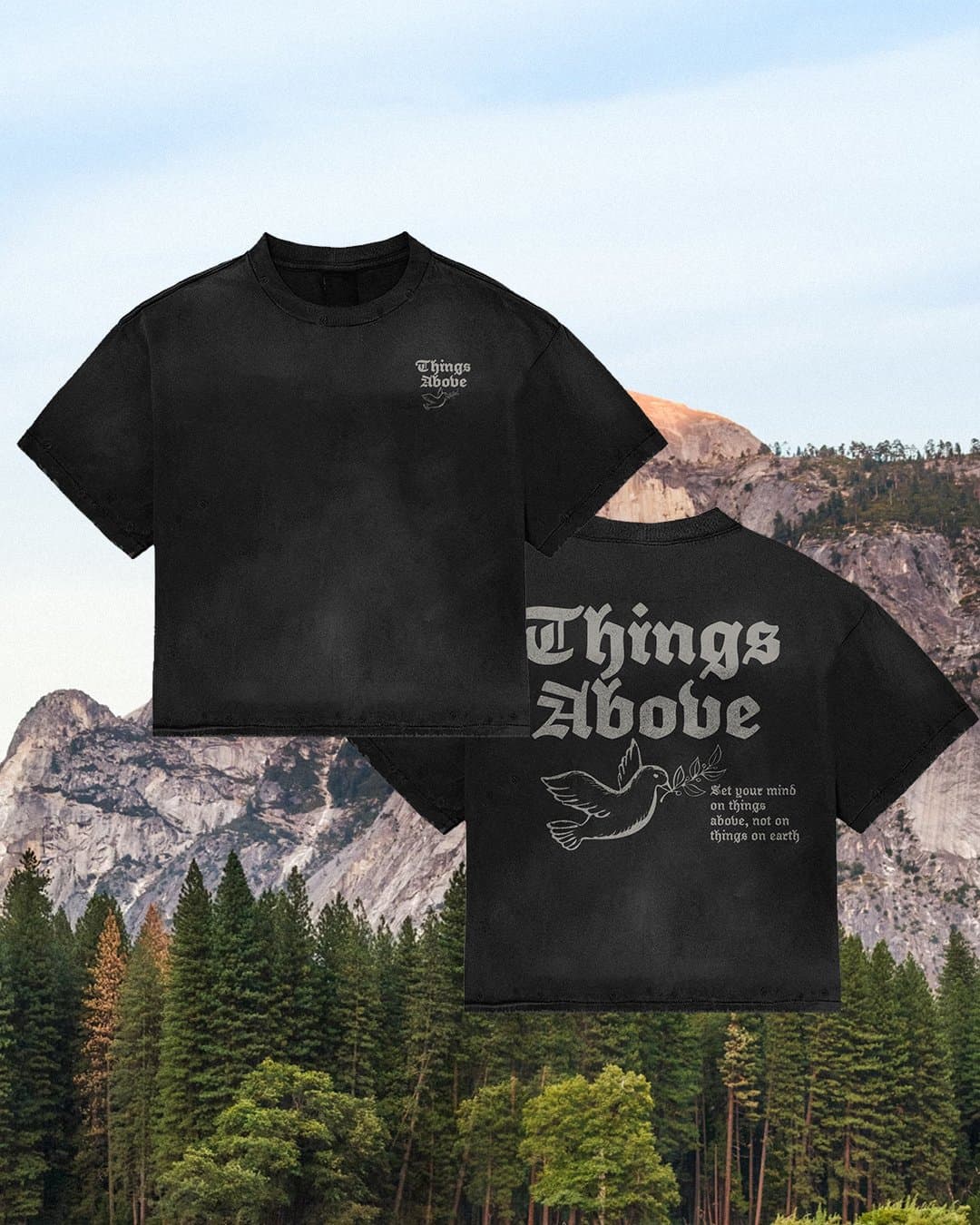 A merch mockup of two washed black tees over a mountain and pine forest, printed Things Above in grey blackletter with a dove holding an olive branch.