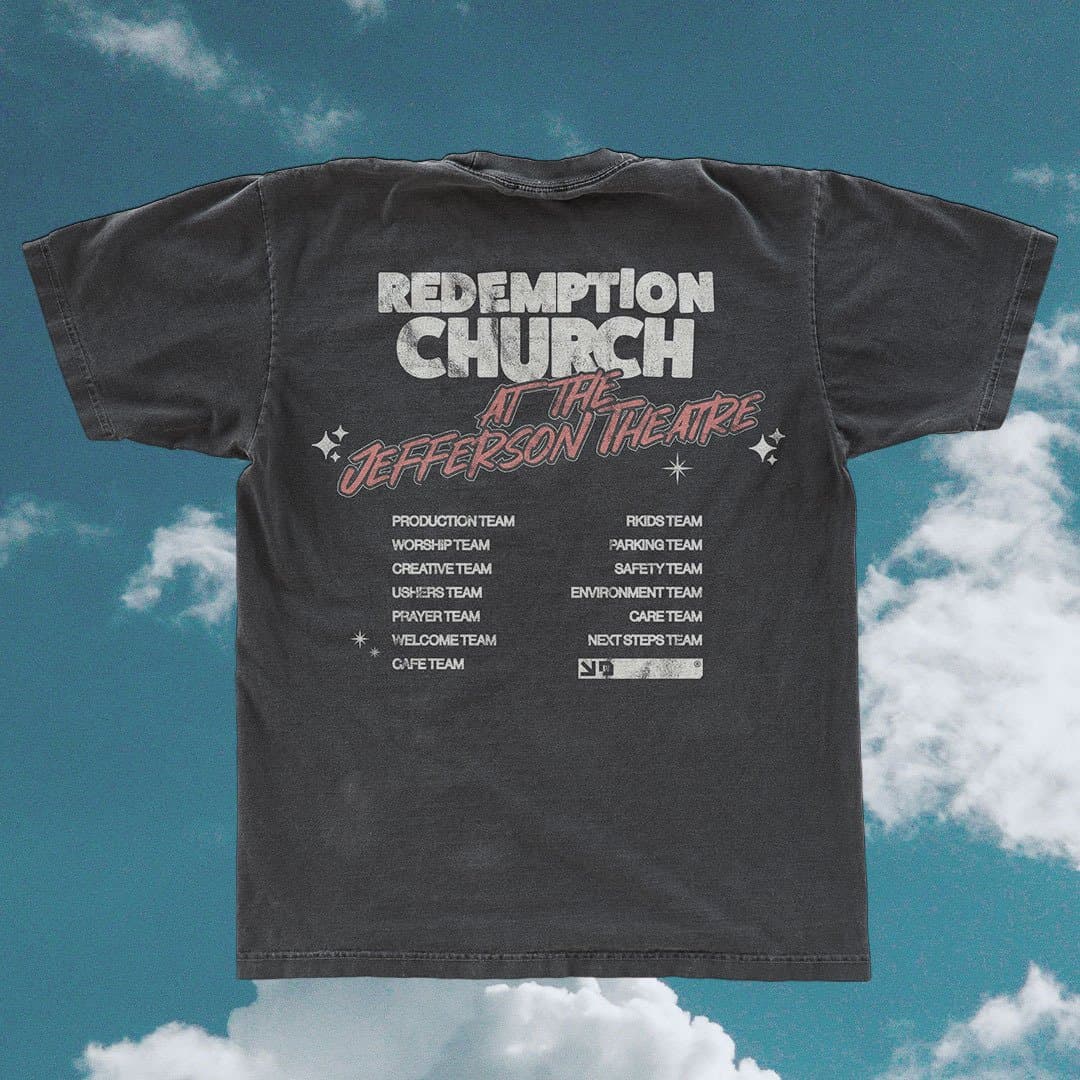 A merch mockup of a washed charcoal tee back against blue sky, reading REDEMPTION CHURCH AT THE Jefferson Theatre in pink script above two columns of volunteer team names.