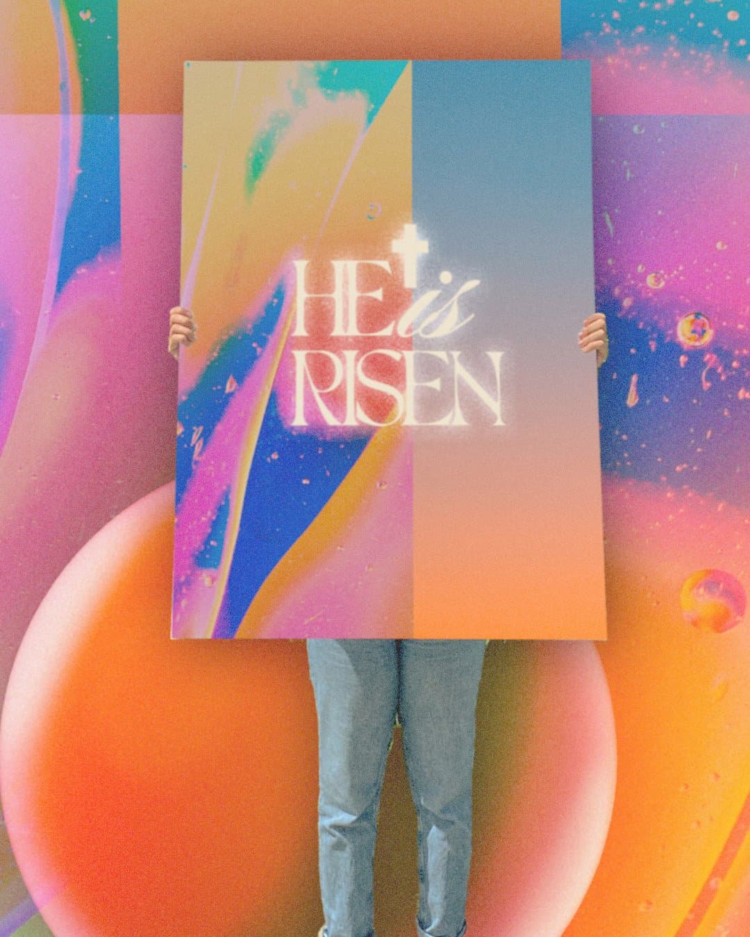 A person holding a poster over their face reading HE is RISEN in glowing cream serif with a cross above, on a split orange and blue gradient, against a matching abstract backdrop.