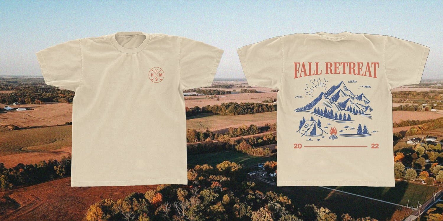 A merch mockup of a cream t shirt front and back reading FALL RETREAT with a blue line drawn mountain, tent and campfire and the year 2022, over an aerial autumn farmland photo.