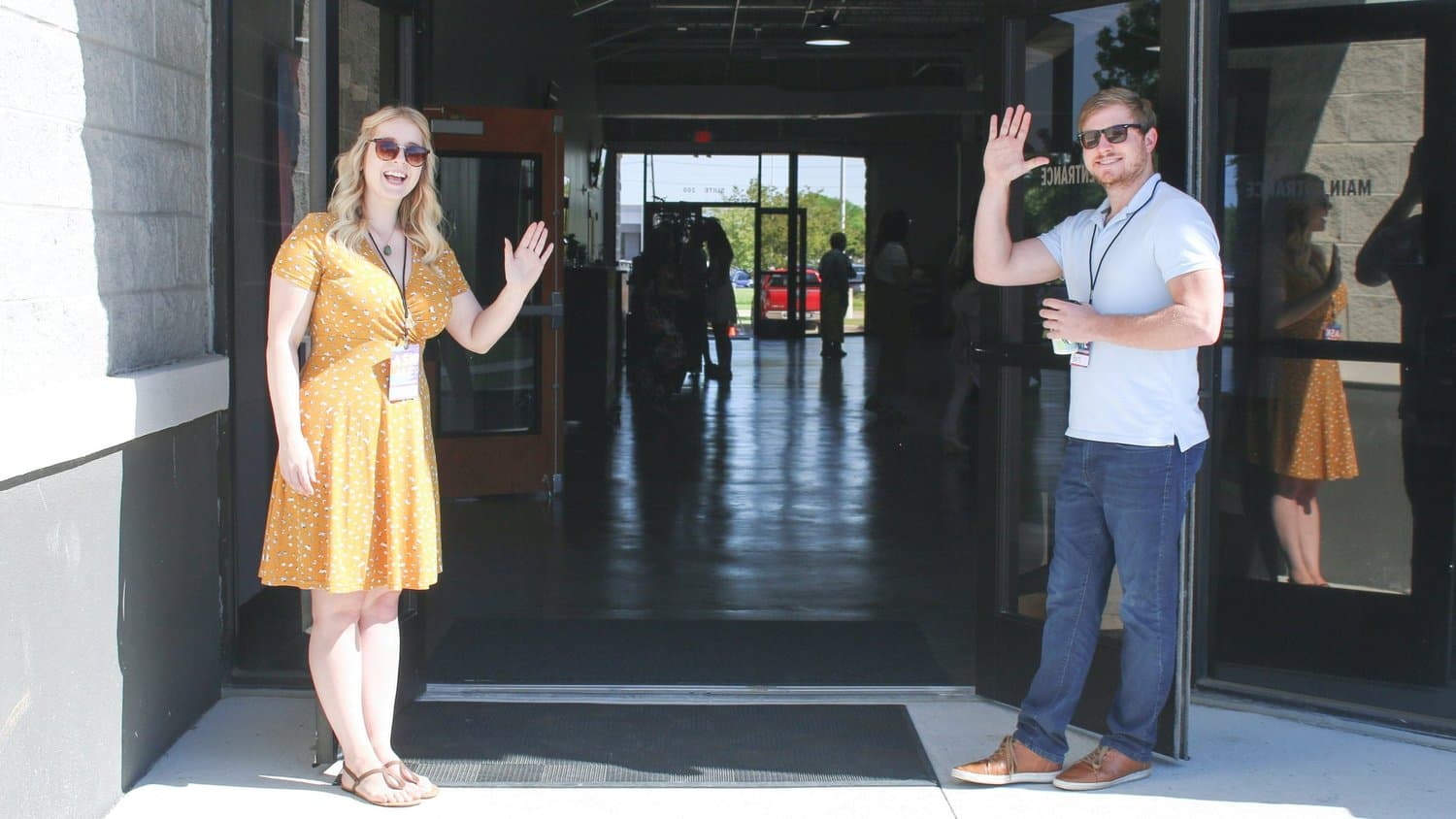 A photo of two lanyard wearing greeters in sunglasses waving from either side of an open glass church entrance on a sunny morning.