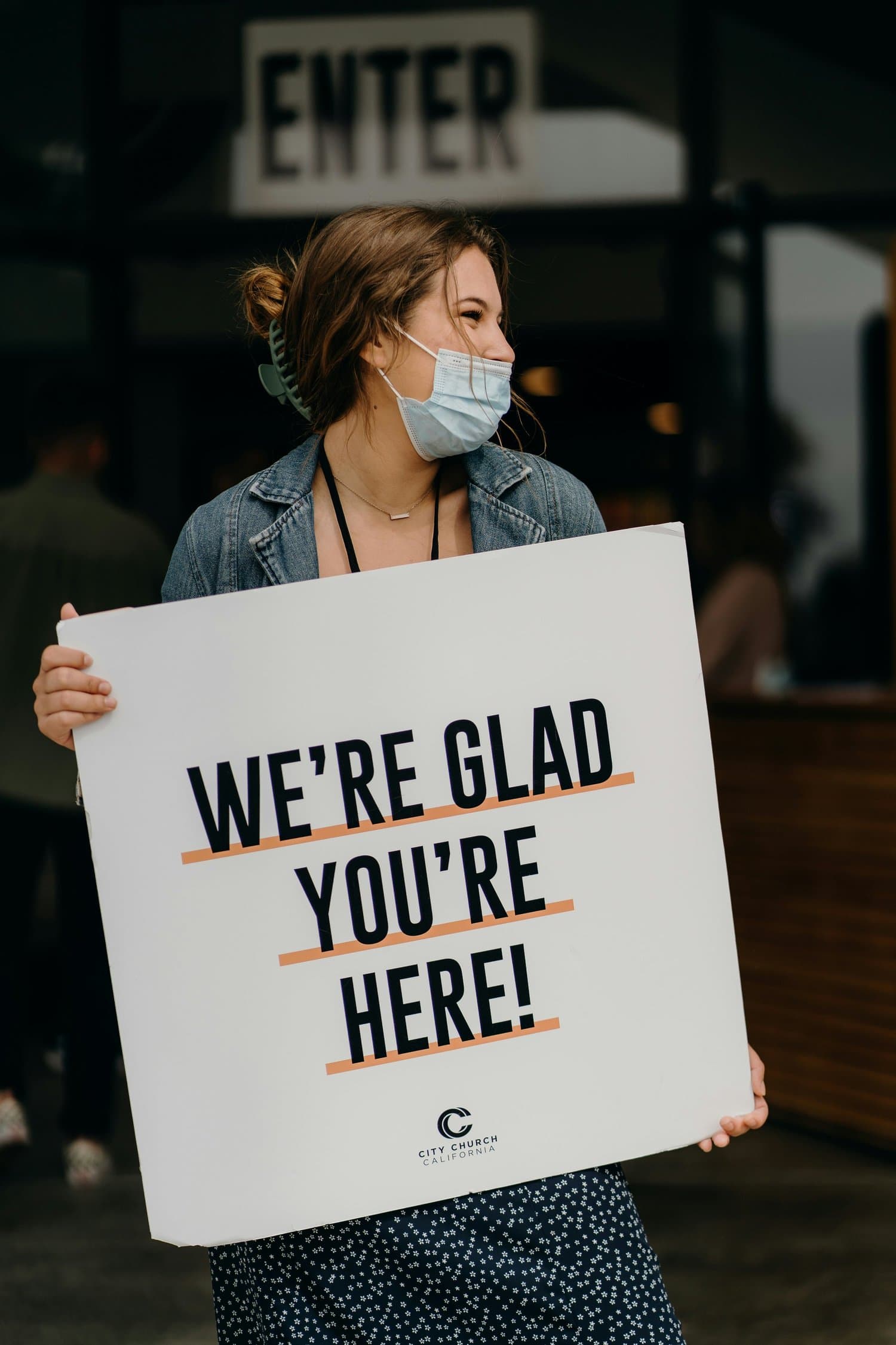 A photo of a volunteer in a denim jacket and face mask holding a large white sign reading WE'RE GLAD YOU'RE HERE! in navy capitals with peach underlines, under an ENTER sign.