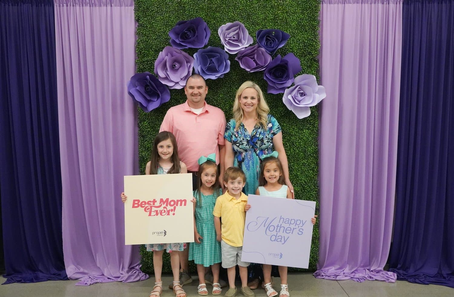 A family of six posed at a Mother's Day photo backdrop of purple drapes and a greenery wall with oversized paper roses, holding Best Mom Ever! and happy Mother's day signs.