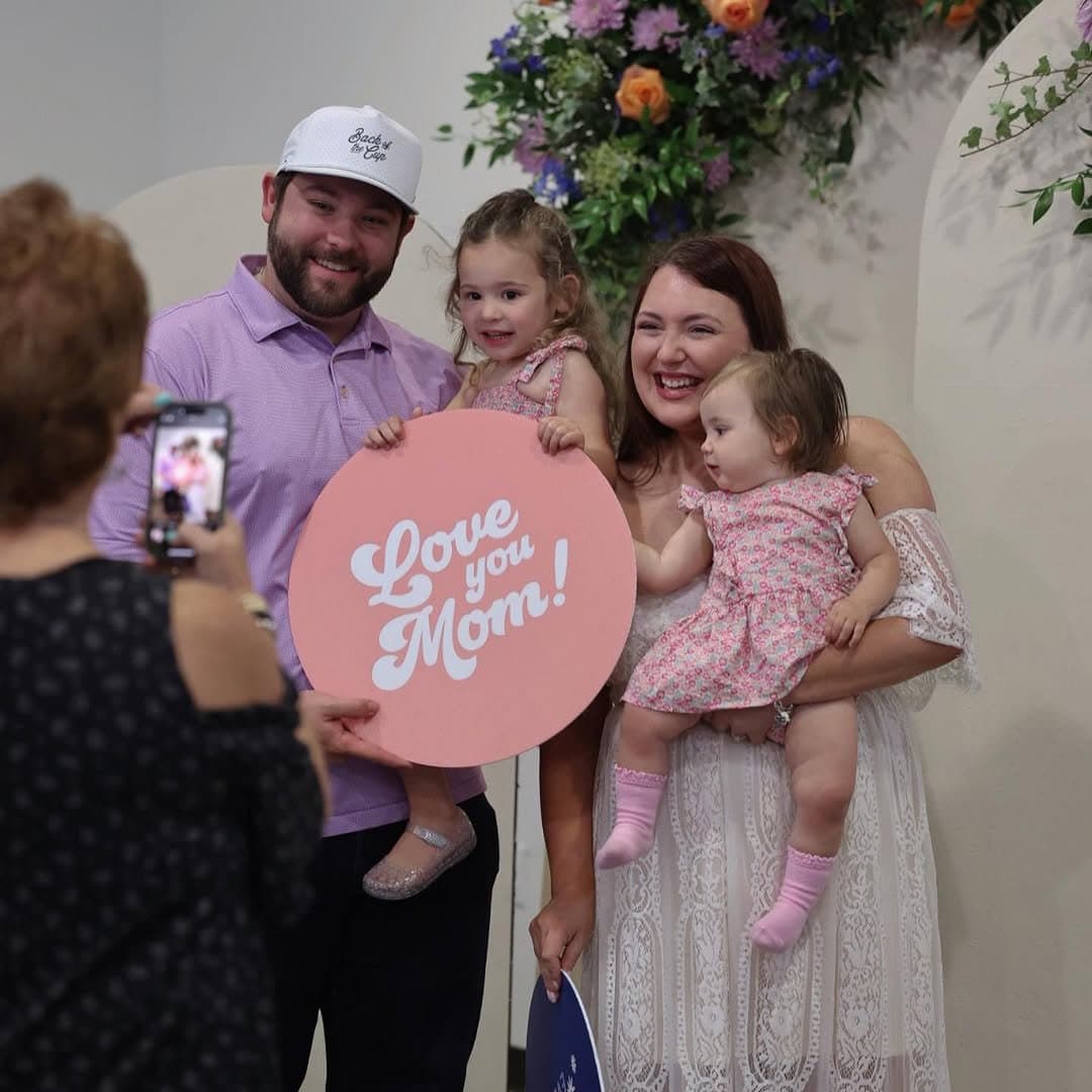 A family photographed at a floral arch holding a round coral sign reading Love you Mom! in white retro script, while a phone captures the shot.