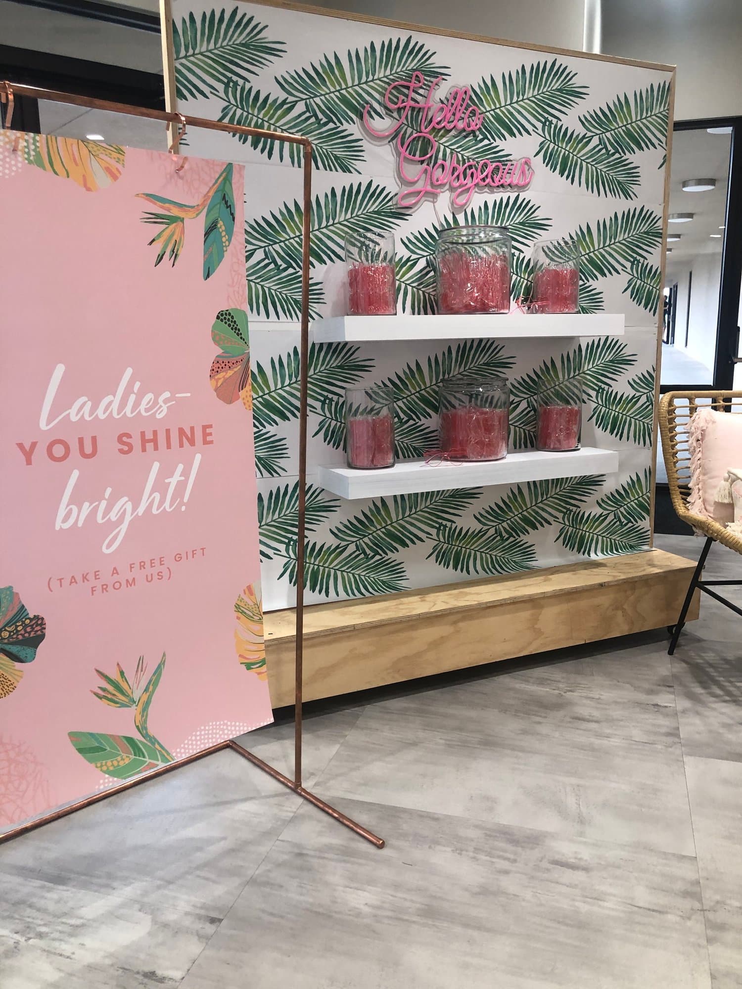 A church lobby gift station with a pink sign reading Ladies, YOU SHINE bright! and TAKE A FREE GIFT FROM US, beside a palm print wall with a pink Hello Gorgeous neon and jars of favours.