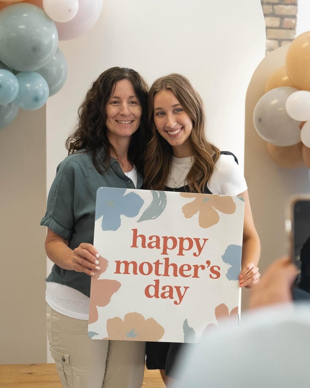 A mother and teenage daughter holding a square happy mother's day sign in rust serif with painted blue and tan blooms, framed by pastel balloon garlands.