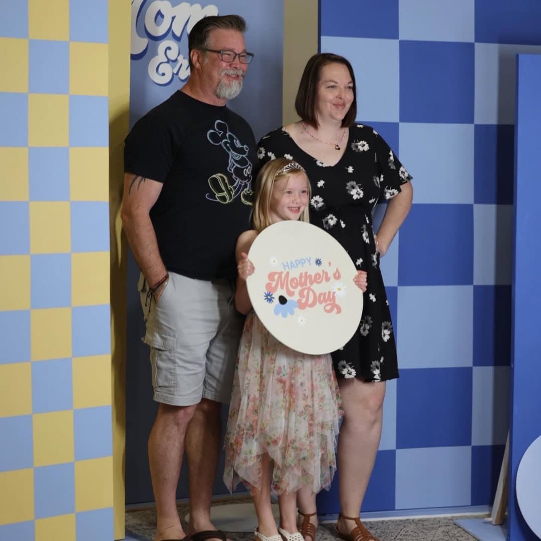 A family posed against a blue and yellow checkerboard photo wall, a girl holding an oval cream sign reading HAPPY Mother's Day in coral script with daisies.