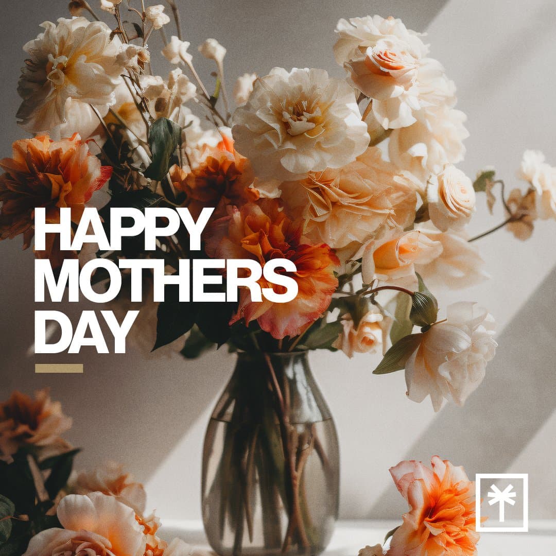 A Mother's Day social post reading HAPPY MOTHERS DAY in white block capitals over a sunlit photo of cream and orange roses in a glass vase, with a small square cross mark logo.