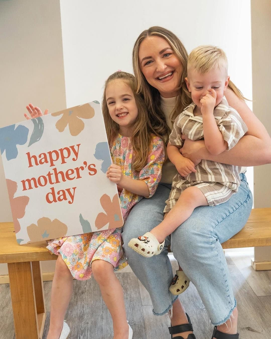 A photo booth moment with a mom and two young children on a bench, holding a cream sign reading happy mother's day in rust serif among painted blue and tan flowers.