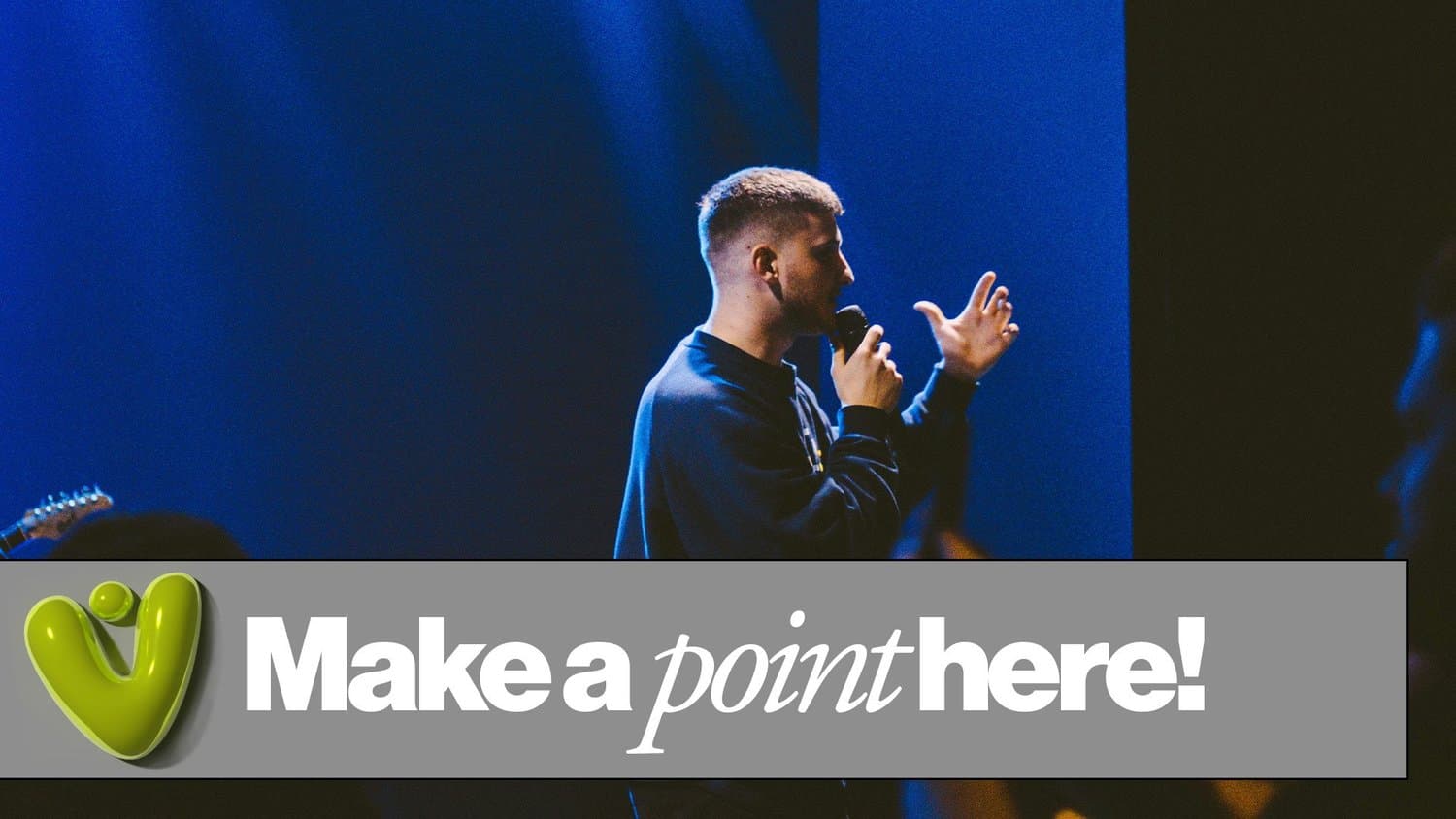 A lower third bar over a stage photo of a young man speaking under blue light, reading Make a point here! in white sans and italic serif beside a glossy green heart logo.