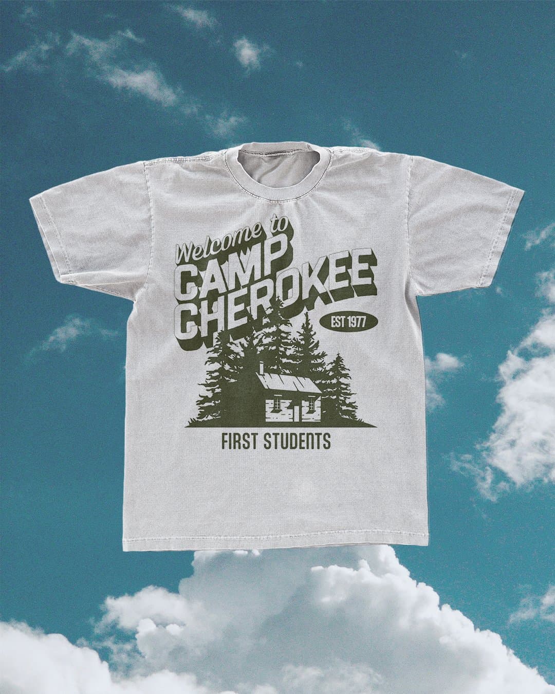 A merch mockup of a washed grey tee printed Welcome to CAMP CHEROKEE in olive green with a cabin among pine trees, EST 1977 and FIRST STUDENTS, floating against blue sky.