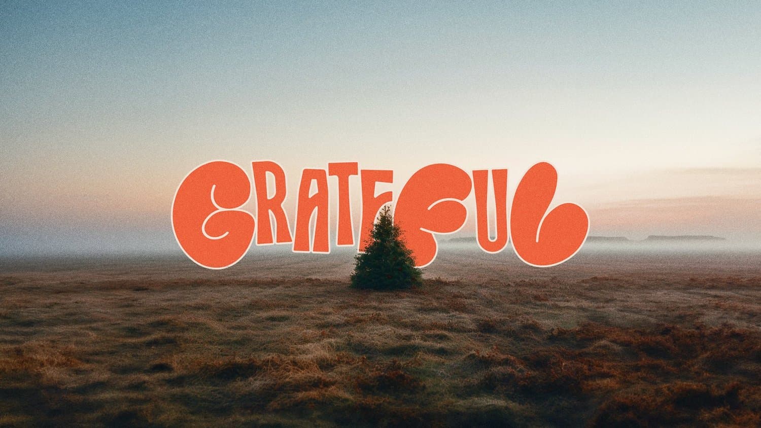 A Thanksgiving slide reading GRATEFUL in orange bubble letters, over a hazy sunrise photo of a lone evergreen in an open field.