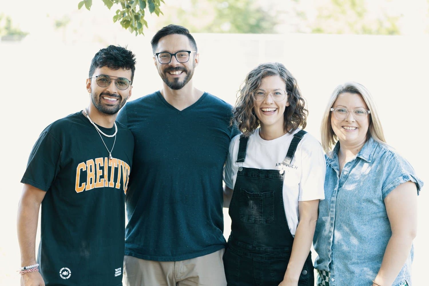 Four smiling team members standing outdoors in daylight, one wearing a black tee printed CREATIVE in collegiate lettering.